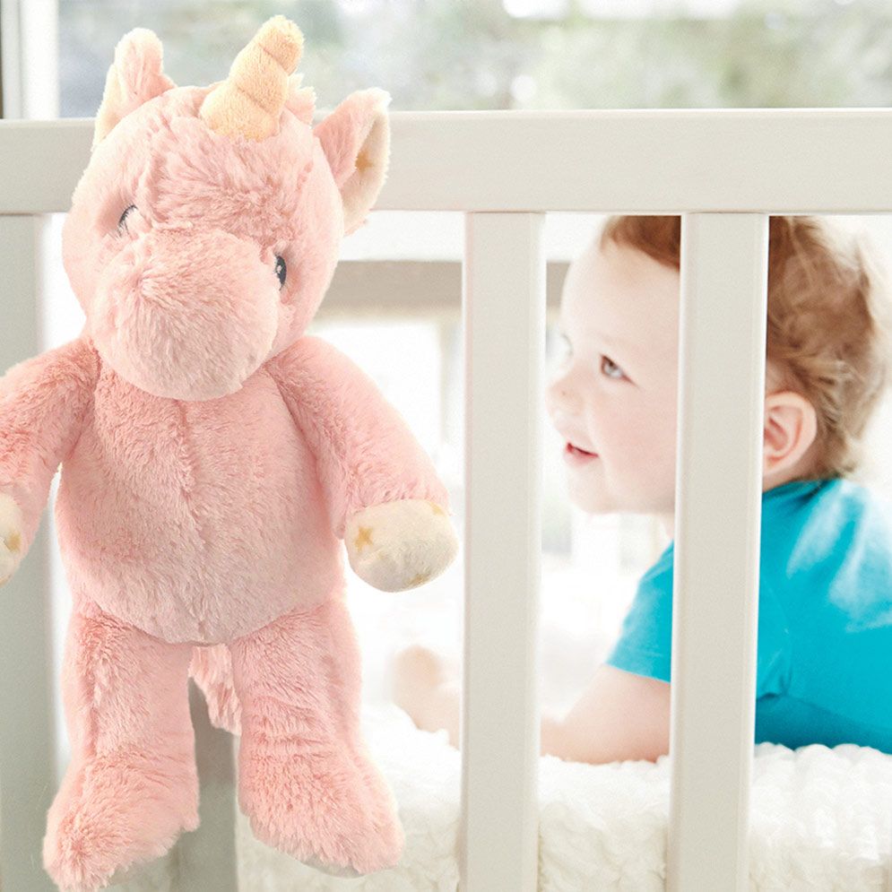 Cloud b sleep aid Ella unicorn, lifestyle photo of pink plush toy hanging on crib rail, baby in background