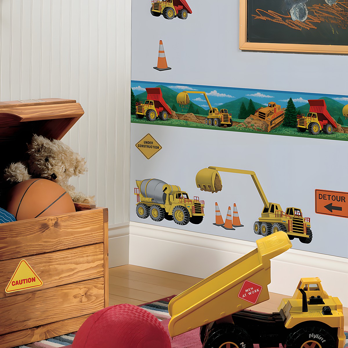 RoomMates Bordüre Under Construction, illustrierter Baustellen-Bordürenstreifen mit Baggern und Lastwagen über Baustellenfahrzeug-Wandstickern an einer hellblauen Kinderzimmerwand