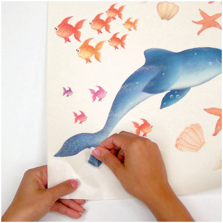 RoomMates wall decal Mermaid, hands peeling the dolphin motif with mermaid off the backing sheet, surrounded by fish and shells