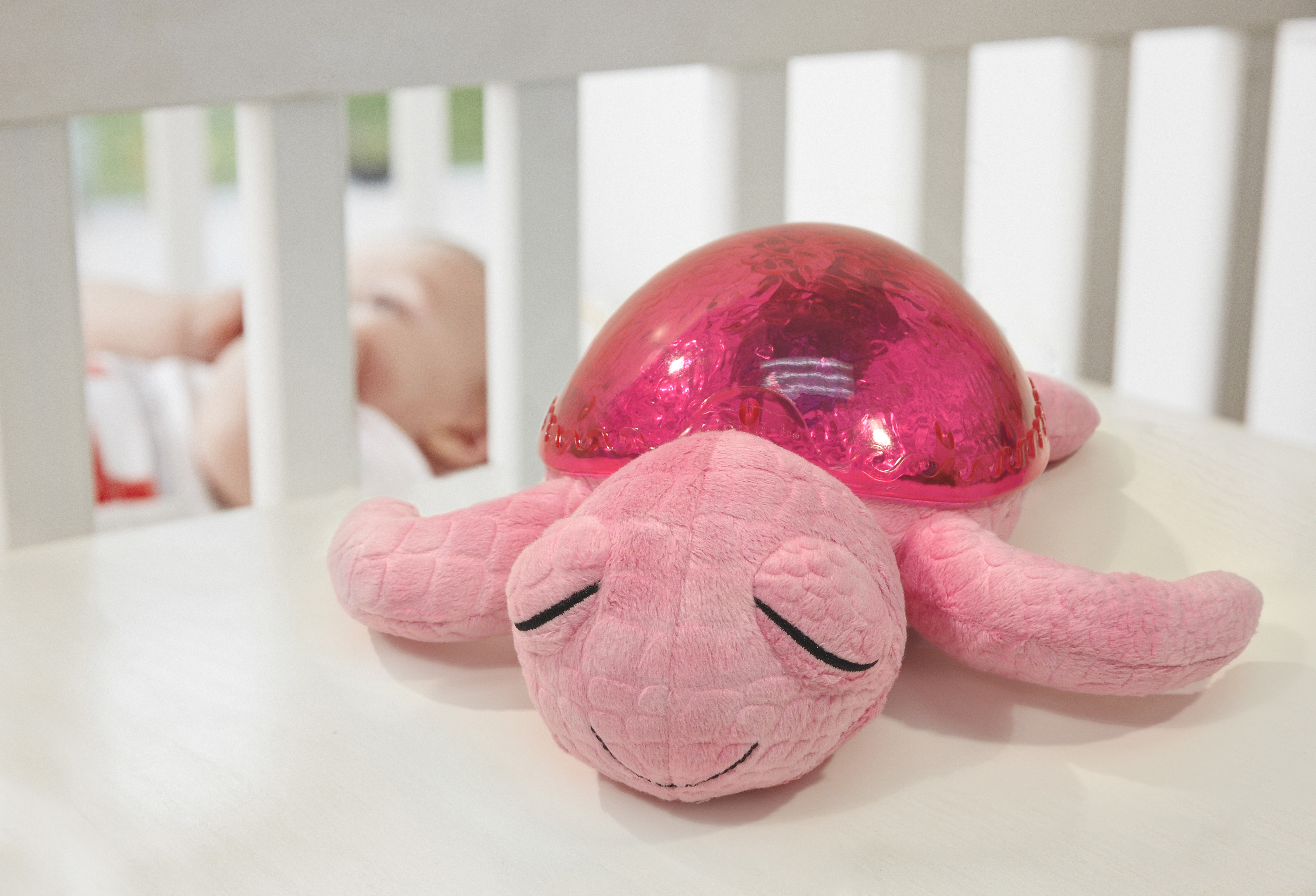 Cloud b sleep aid Tranquil Turtle Pink, plush toy on crib rail with sleeping baby in the background